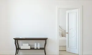 Un corridoio con un tavolino scuro e una porta aperta che rivela una stanza da letto. La porta interna è dipinta di bianco, con un design classico.