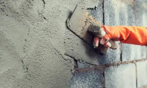 Mano con guanto arancione stende malta da costruzione su un muro di blocchi di cemento con una cazzuola.