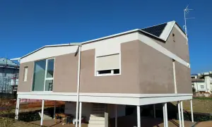 Casa moderna con intonaco argilla, pannelli solari e ampie vetrate. La struttura è rialzata su pilastri bianchi.
