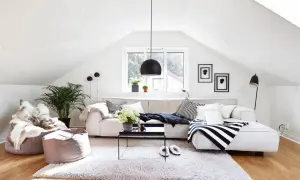 Arredare mansarda: un salotto accogliente con divano angolare, pouf, tappeto soffice e piante. Luce naturale entra dalla finestra.
