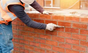 Muratore con guanti e cazzuola stende malta modellabile tra i mattoni rossi di un muro in costruzione.