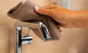 Mano che pulisce un rubinetto cromato con un panno marrone, mostrando come togliere il calcare dai rubinetti per renderli brillanti.