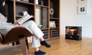 Donna legge un libro seduta su una poltrona, mentre un fungo a cherosene scalda l'ambiente. Una casa senza riscaldamento, ma con un tocco di calore.