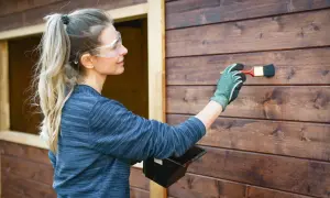 Donna con occhiali protettivi applica vernice su una parete di legno scuro. Si può verniciare sopra l'impregnante per un nuovo look.