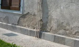 Muro esterno che si sbriciola, mostrando crepe e intonaco scrostato vicino a tubi verticali e una finestra con persiane.