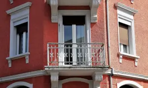 Dettaglio di un muro esterno con finitura a rasatura, finestre con persiane e un piccolo balcone con ringhiera in ferro battuto.