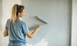 Donna con i capelli biondi in coda di cavallo, jeans e maglietta blu, impara come pitturare con il rullo una parete grigia.