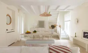 Un soggiorno luminoso con soffitto a travi e **bianco caldo pareti**. Tavolo da pranzo in legno, sedie, panca e una scala bianca conducono a un piano superiore.
