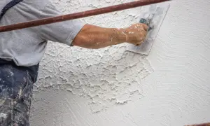 Mano di un operaio che applica intonaco rustico con una spatola, creando una texture grezza e irregolare su una parete.