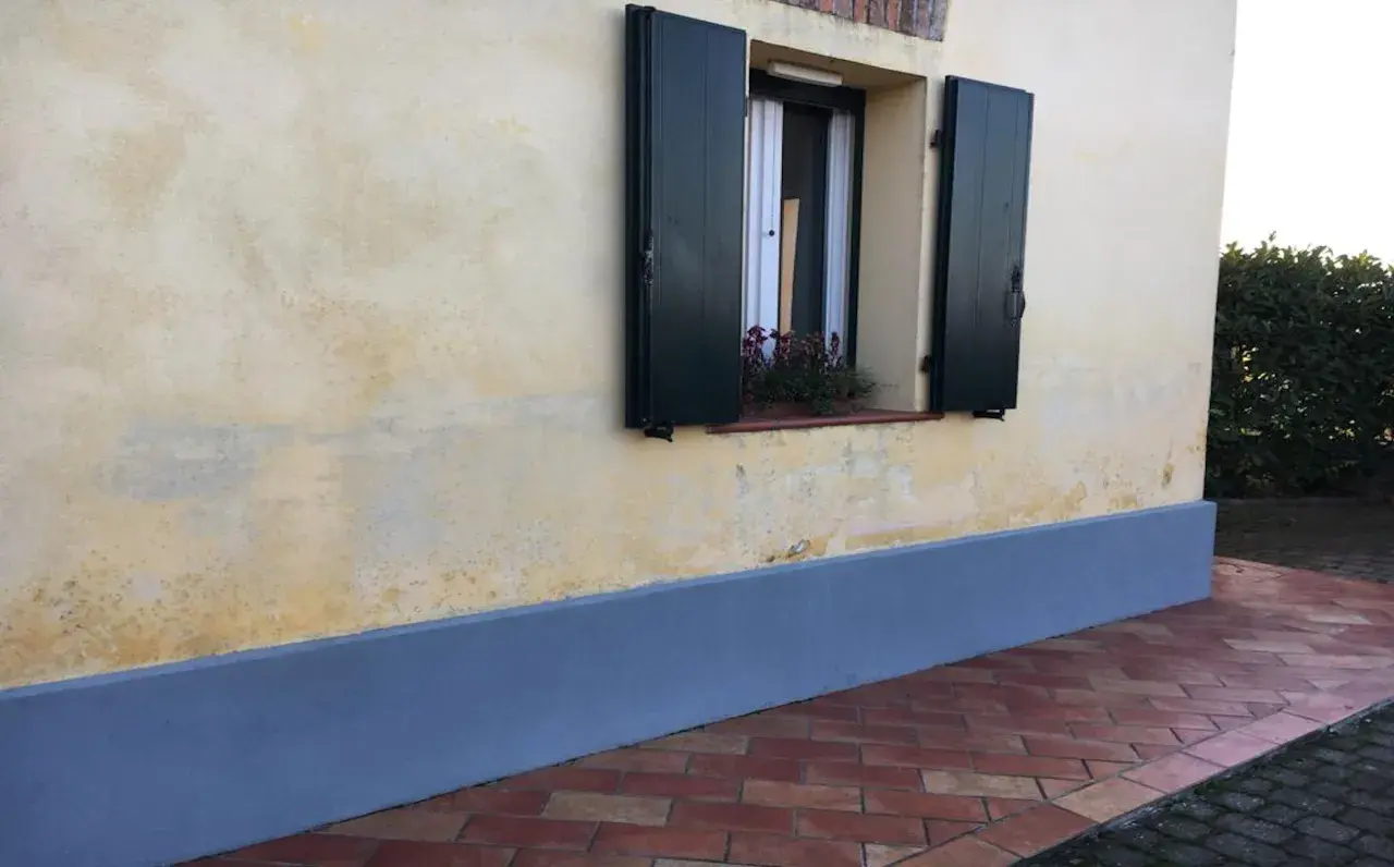 Muro con rinzaffo storico, una finestra con persiane verdi e un davanzale fiorito. Pavimentazione in cotto.