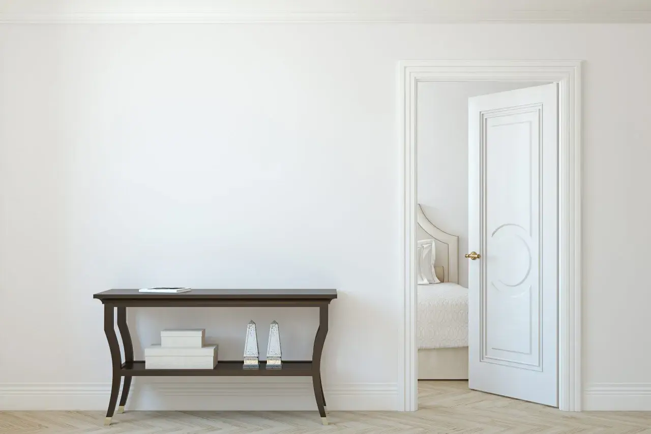 Un corridoio con un tavolino scuro e una porta aperta che rivela una stanza da letto. La porta interna è dipinta di bianco, con un design classico.