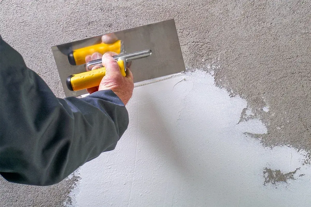 Mano con spatola gialla applica pittura rasante su muro grezzo, creando una superficie liscia e uniforme.