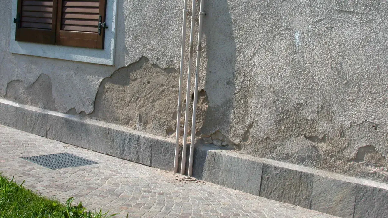 Muro esterno che si sbriciola, mostrando crepe e intonaco scrostato vicino a tubi verticali e una finestra con persiane.