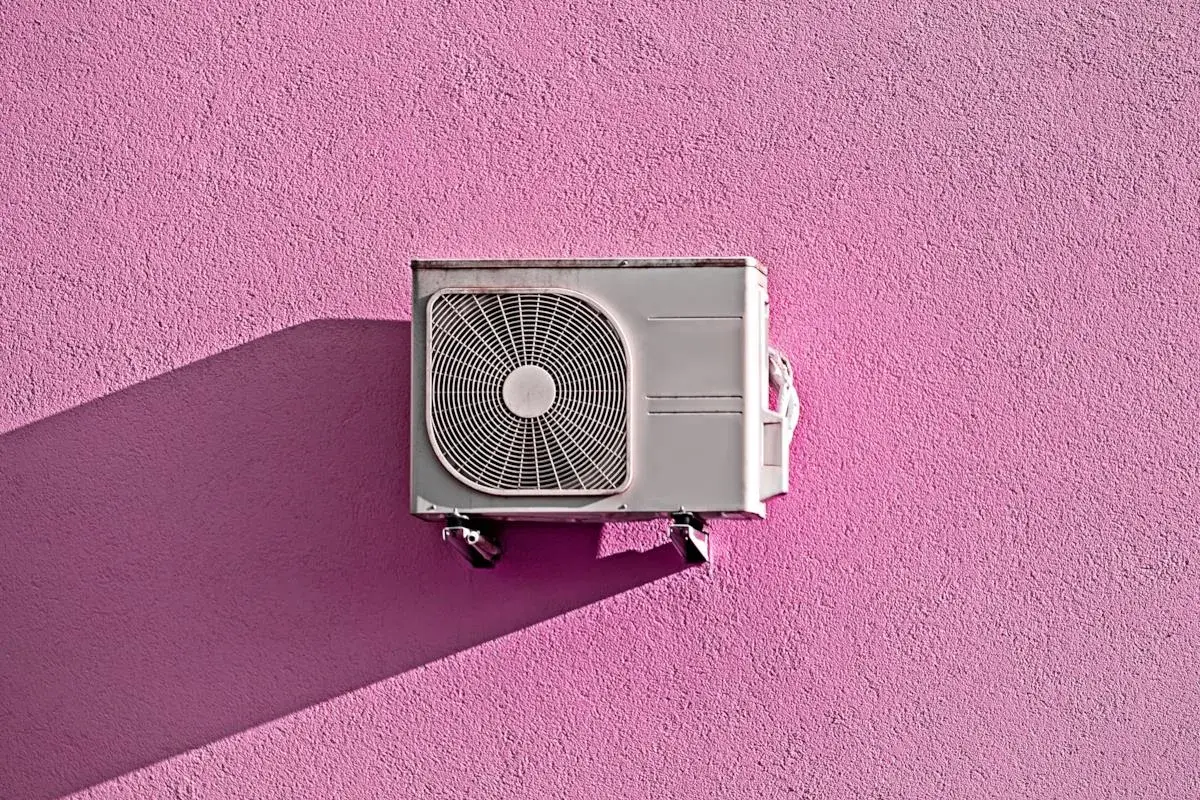 Unità esterna di condizionatore appesa a un muro rosa acceso, con ombra proiettata. Pensando allo smaltimento condizionatori.
