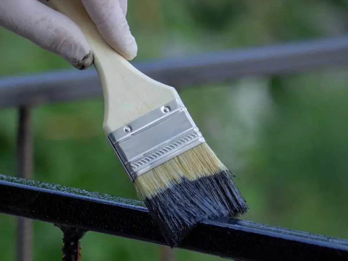 Mano guantata che inizia a verniciare ringhiera con pennello nero.