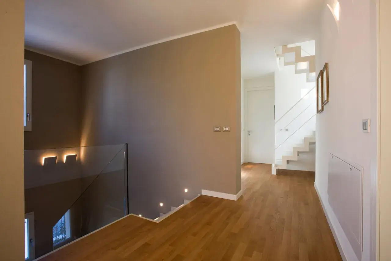Un moderno disimpegno casa con scala bianca a vista, parapetto in vetro e luci a led che illuminano il corridoio.
