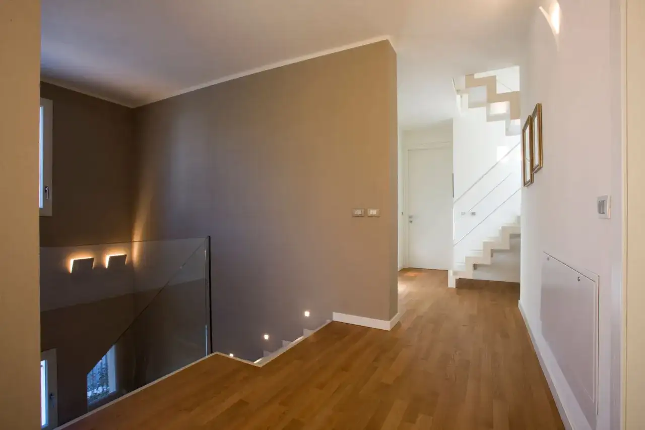 Un moderno disimpegno casa con scala bianca a vista, parapetto in vetro e luci a led che illuminano il corridoio.