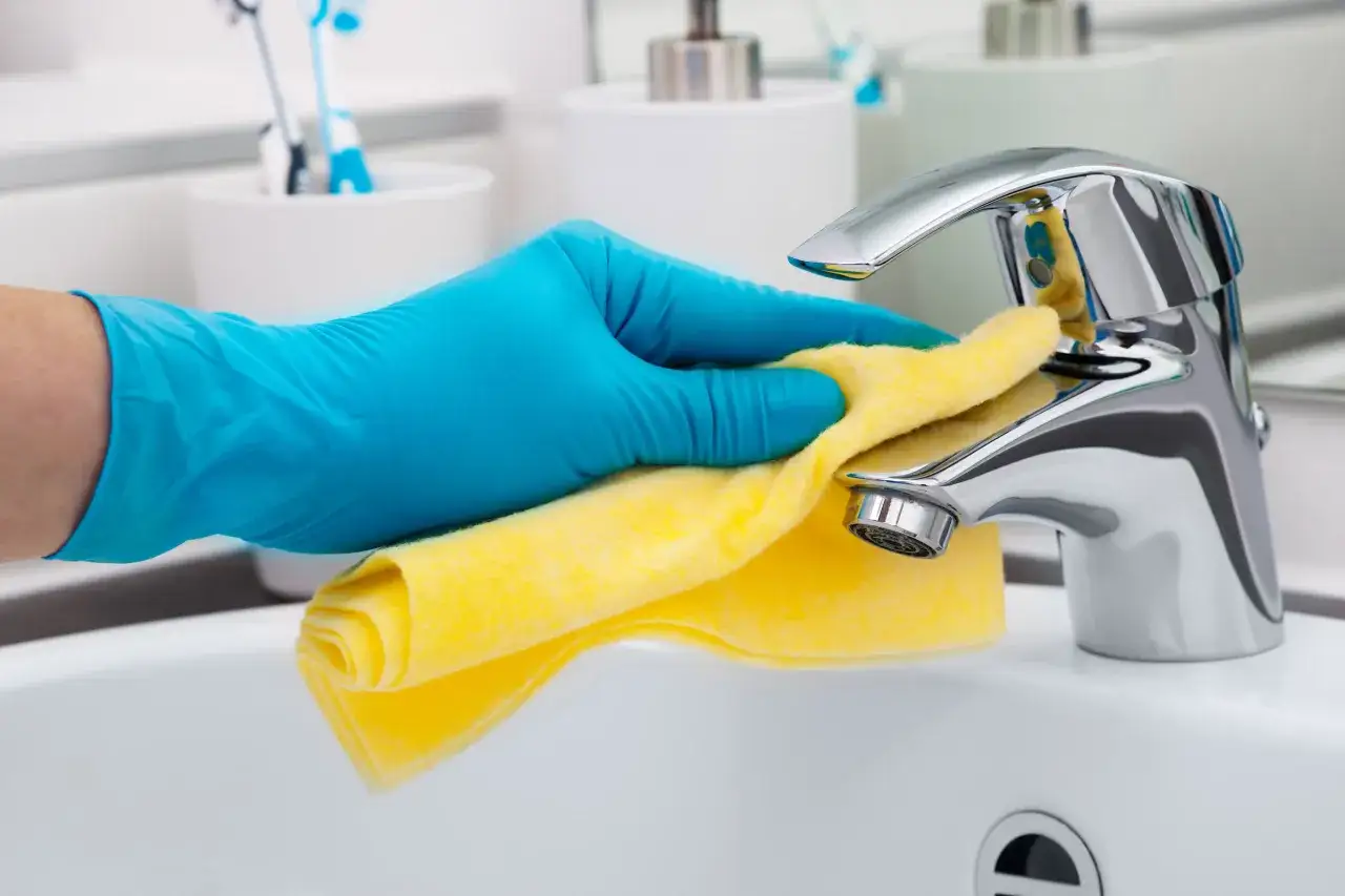 Mano con guanto blu pulisce rubinetti cromati con panno giallo, eliminando il calcare verde.