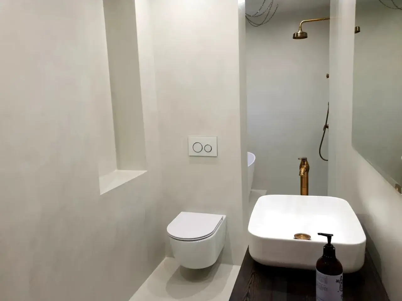 Bagno moderno con pareti in microcemento, lavabo squadrato, doccia a vista e WC sospeso. Finiture in ottone per rubinetteria e soffione doccia.