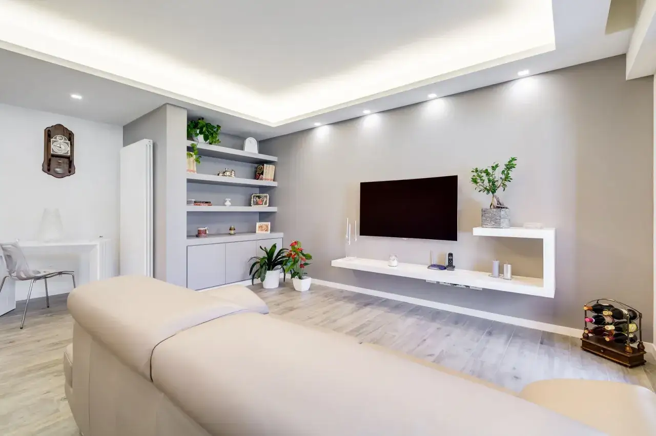 Soggiorno moderno con pareti in cartongesso, un divano chiaro e un mobile TV sospeso. L'open space è illuminato da faretti a soffitto.