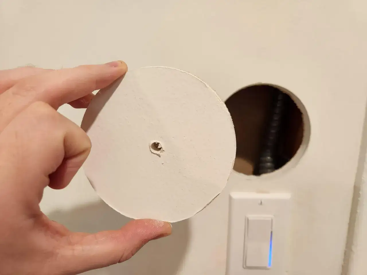 Mano tiene un disco bianco per capire come riempire un grosso buco nel muro. Vicino, un interruttore con luce blu.