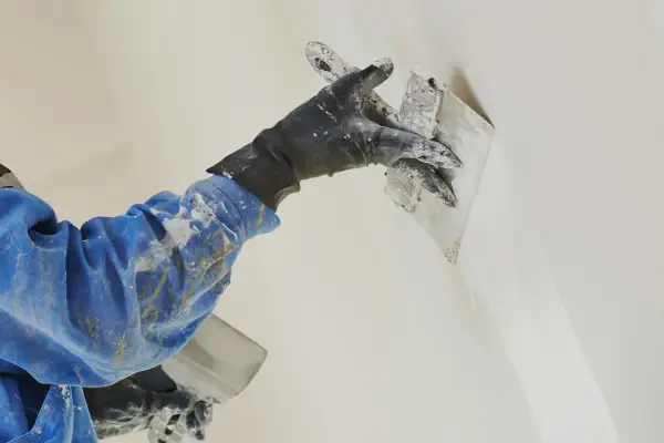 Mano guantata con cazzuola esegue la rasatura di un muro, creando una finitura liscia e uniforme.