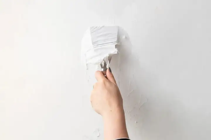 Mano con spatola che rimuove carta da parati bianca da un muro.